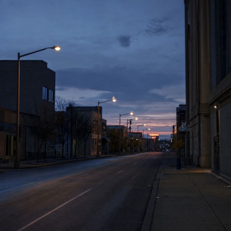 An empty street at dusk leading to a distant lone taqueria sign